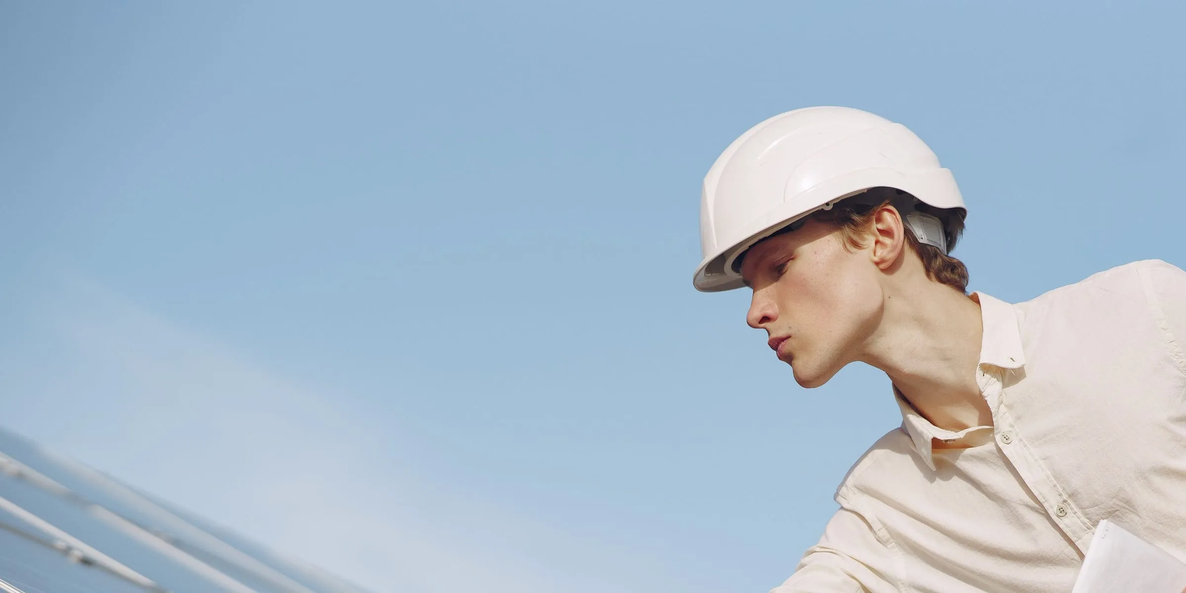 An engineer in a white hard hat checking a solar panel against a clear sky