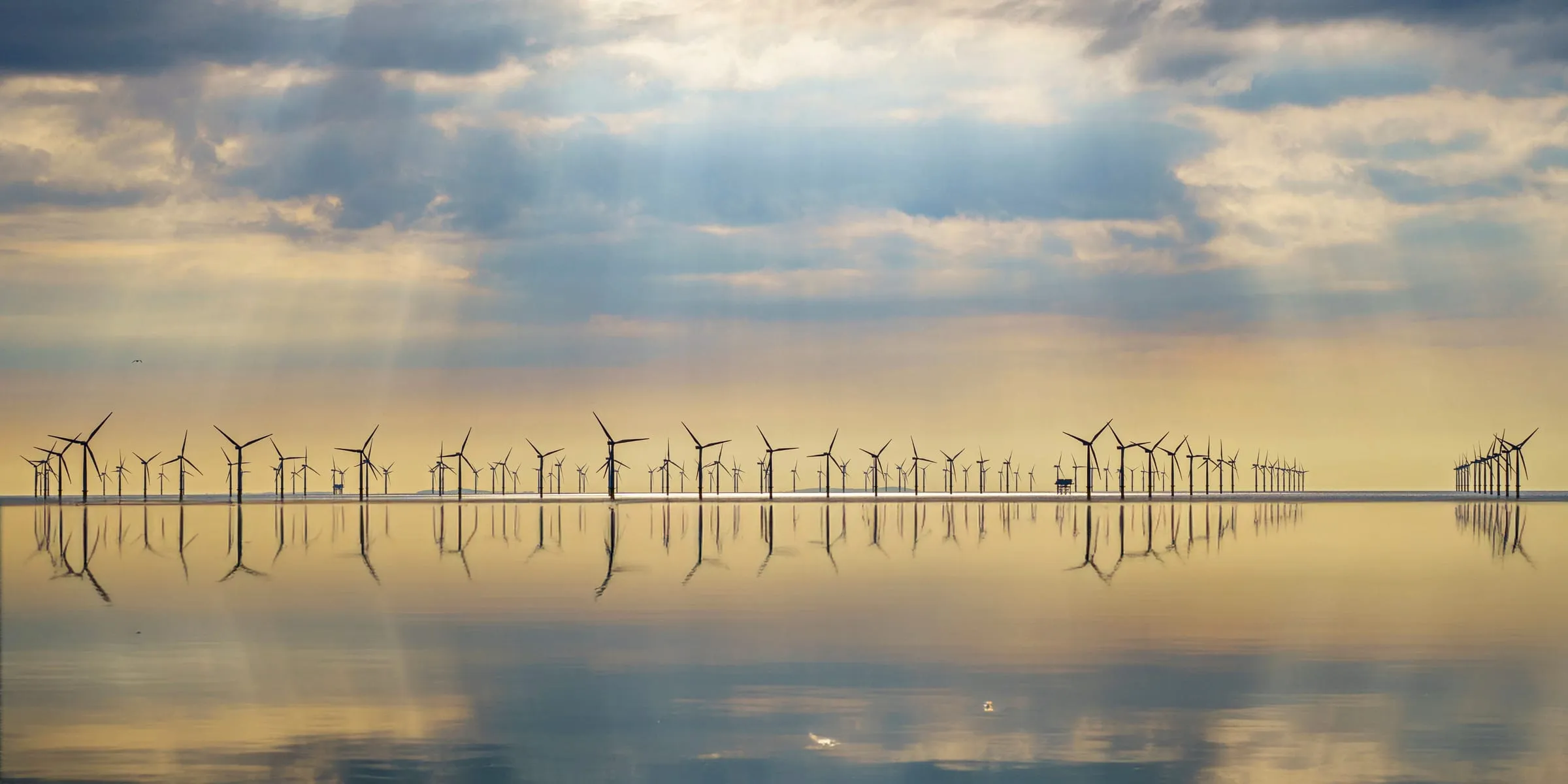 An offshore wind farm on the horizon above still water under a broken sky